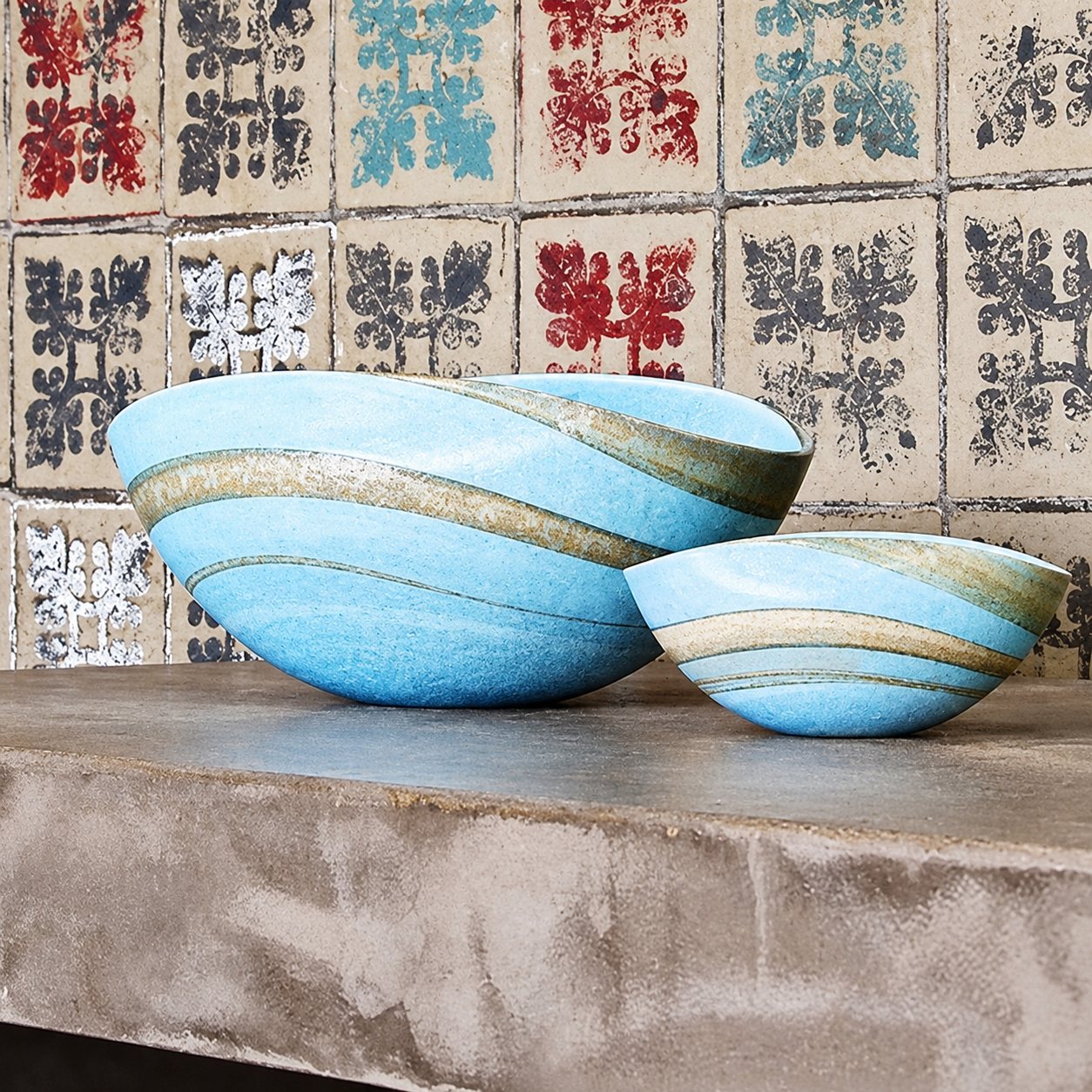 Deux bols en verre soufflé bleu clair à rayures beiges et grises posés sur un plan gris devant un mur décoré de carreaux à motifs.