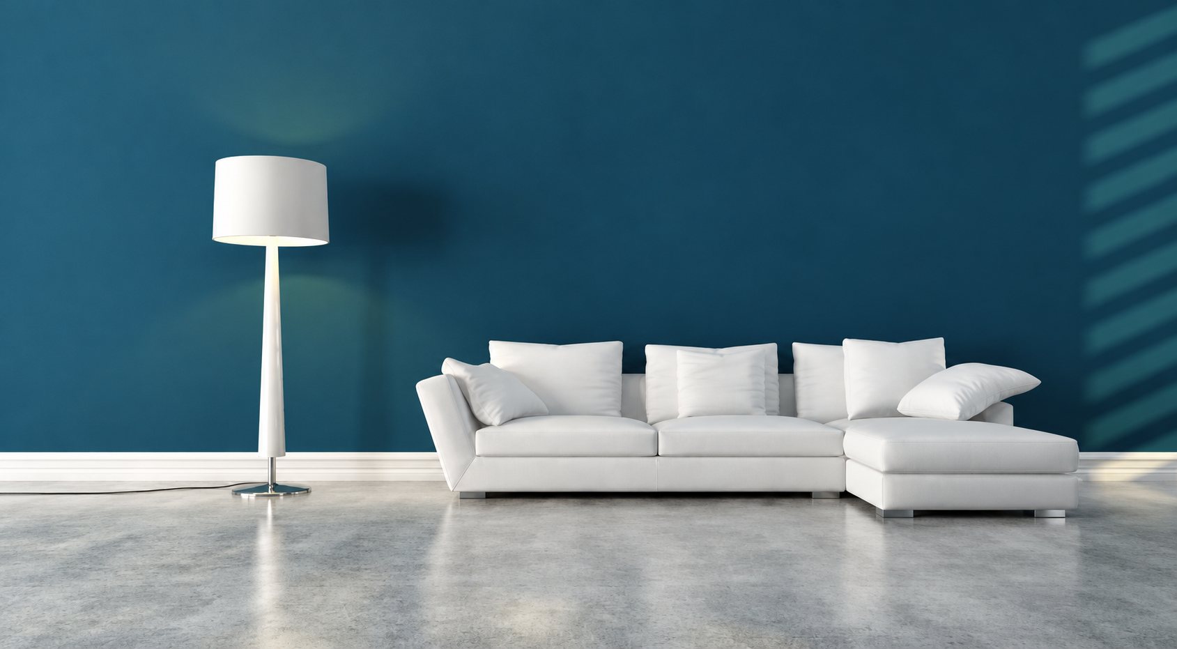 Un canapé d’angle blanc garni de coussins et un lampadaire blanc allumé se trouvent devant un mur bleu foncé, sur un sol gris clair.