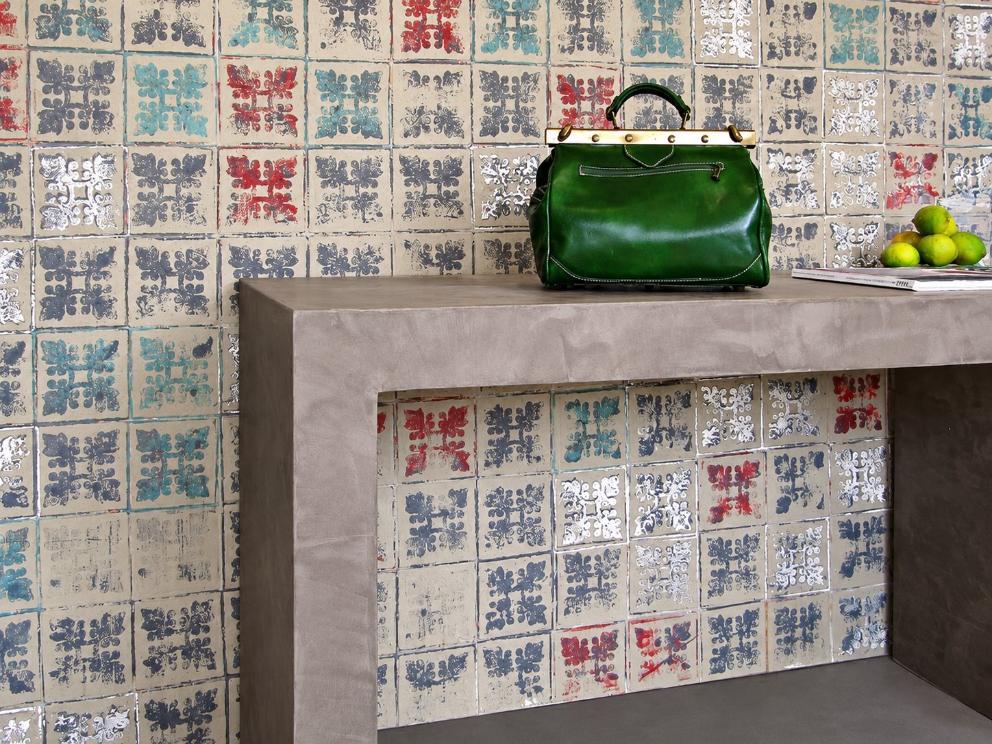 Un sac vert en cuir posé sur une console grise en béton ciré, devant un mur couvert de carreaux à motifs colorés et argentés, avec des citrons verts à droite.