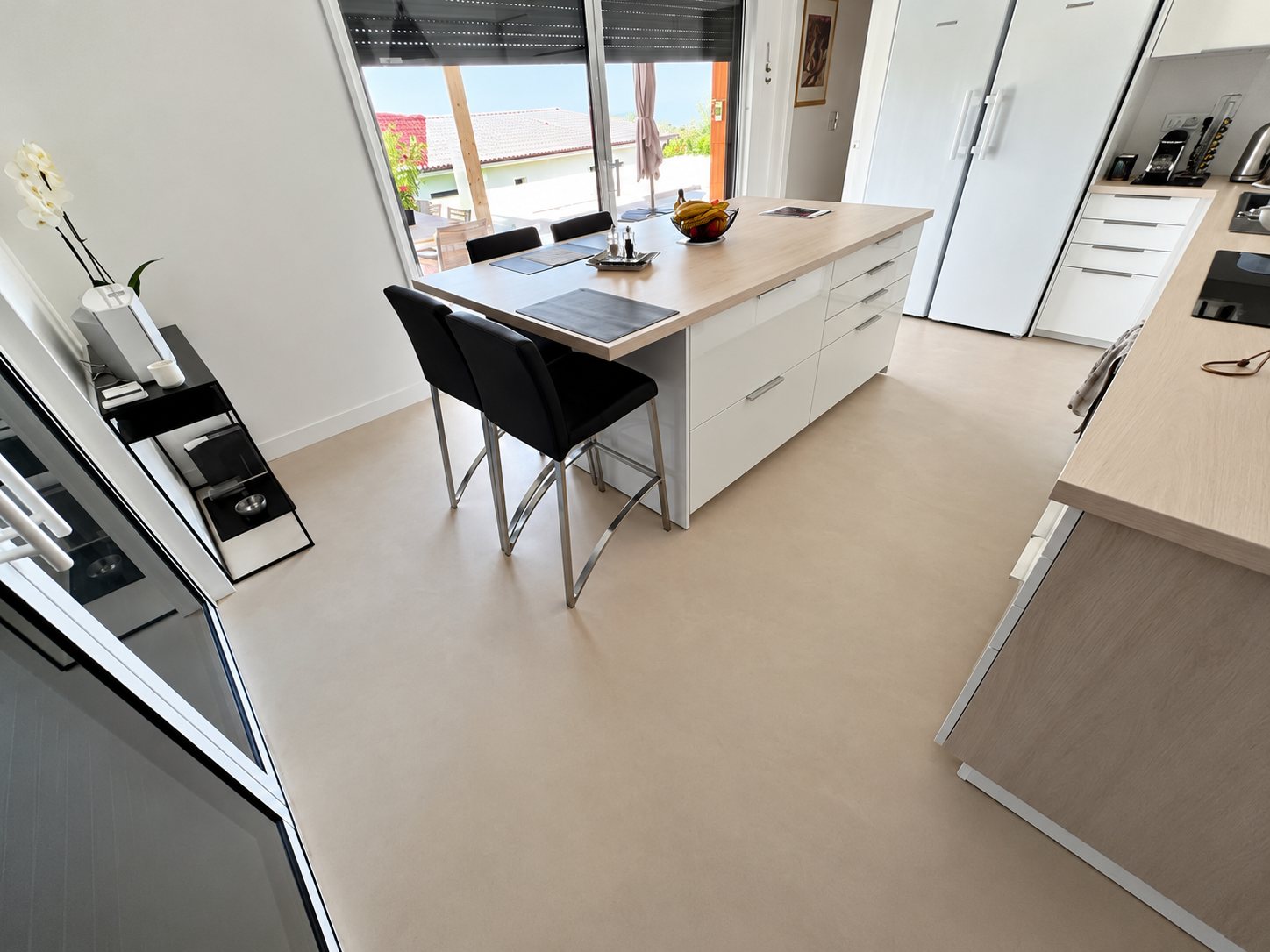 Une cuisine lumineuse avec un îlot central blanc, deux tabourets noirs et un grand sol lisse beige près d’une baie vitrée.