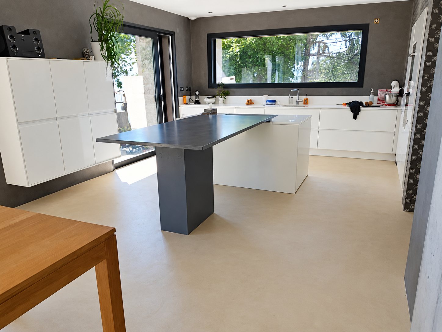Vaste cuisine contemporaine aux meubles blancs laqués, avec un îlot central à plateau gris, un sol clair en béton ciré et une grande fenêtre donnant sur la verdure.