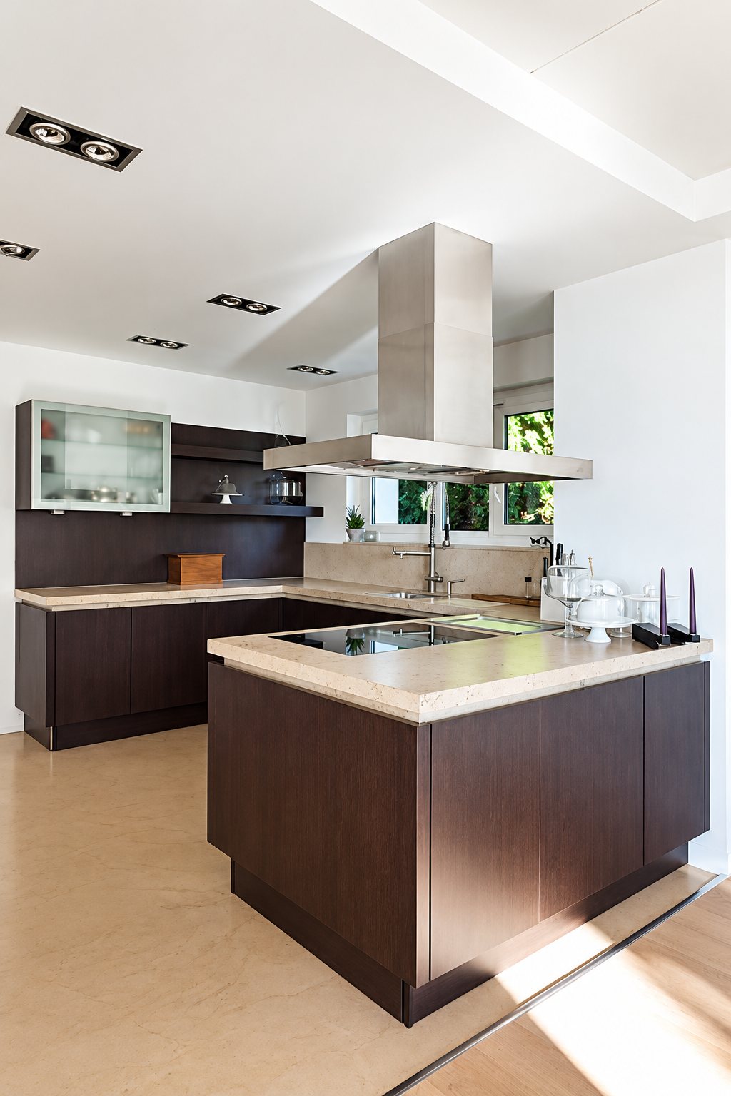Cuisine contemporaine lumineuse avec meubles en bois foncé, plan de travail beige, plaque de cuisson et hotte en inox suspendue.