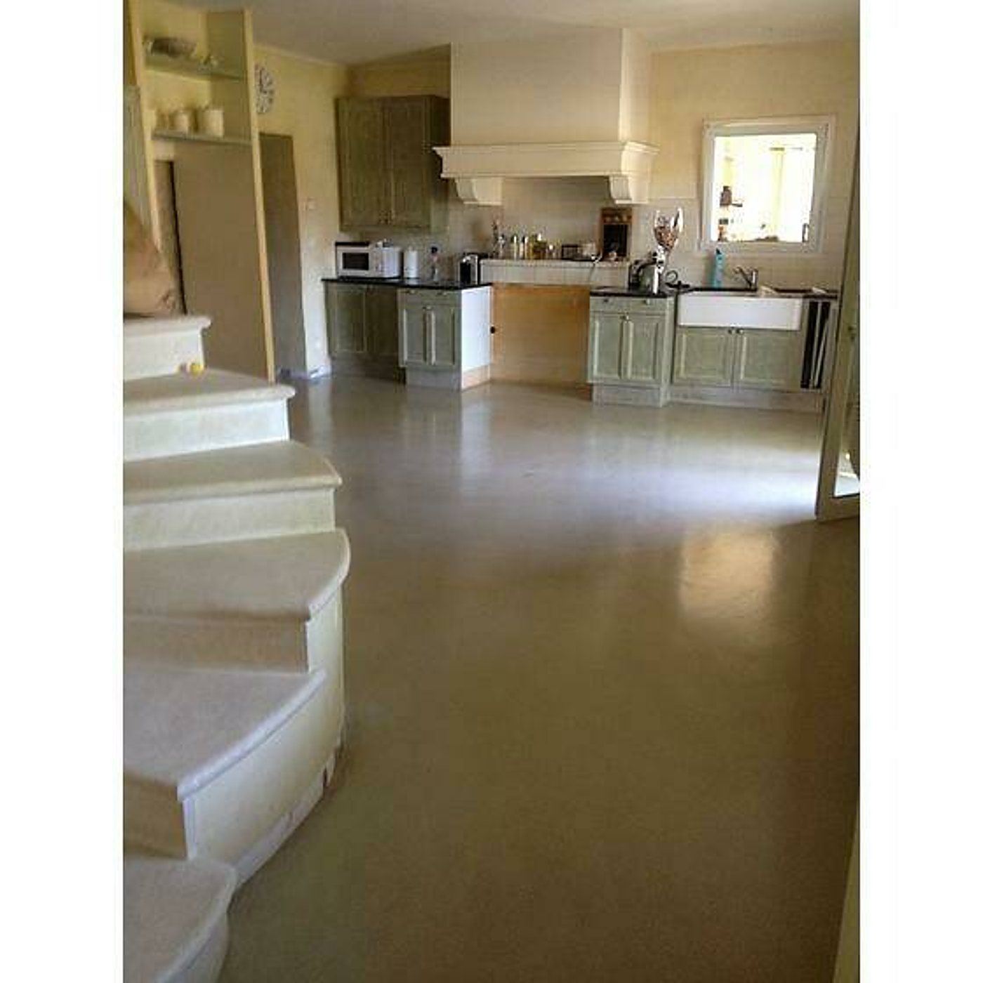 Grande cuisine ouverte aux meubles vert pâle, avec un sol en béton ciré beige lisse et un escalier arrondi au premier plan.