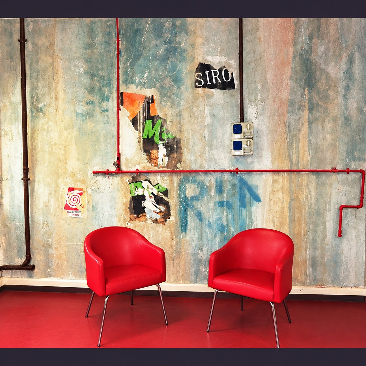 Deux fauteuils rouges au pied d’un mur patiné orné de tuyaux rouges, d’affiches déchirées et de graffitis, sur un sol rouge.