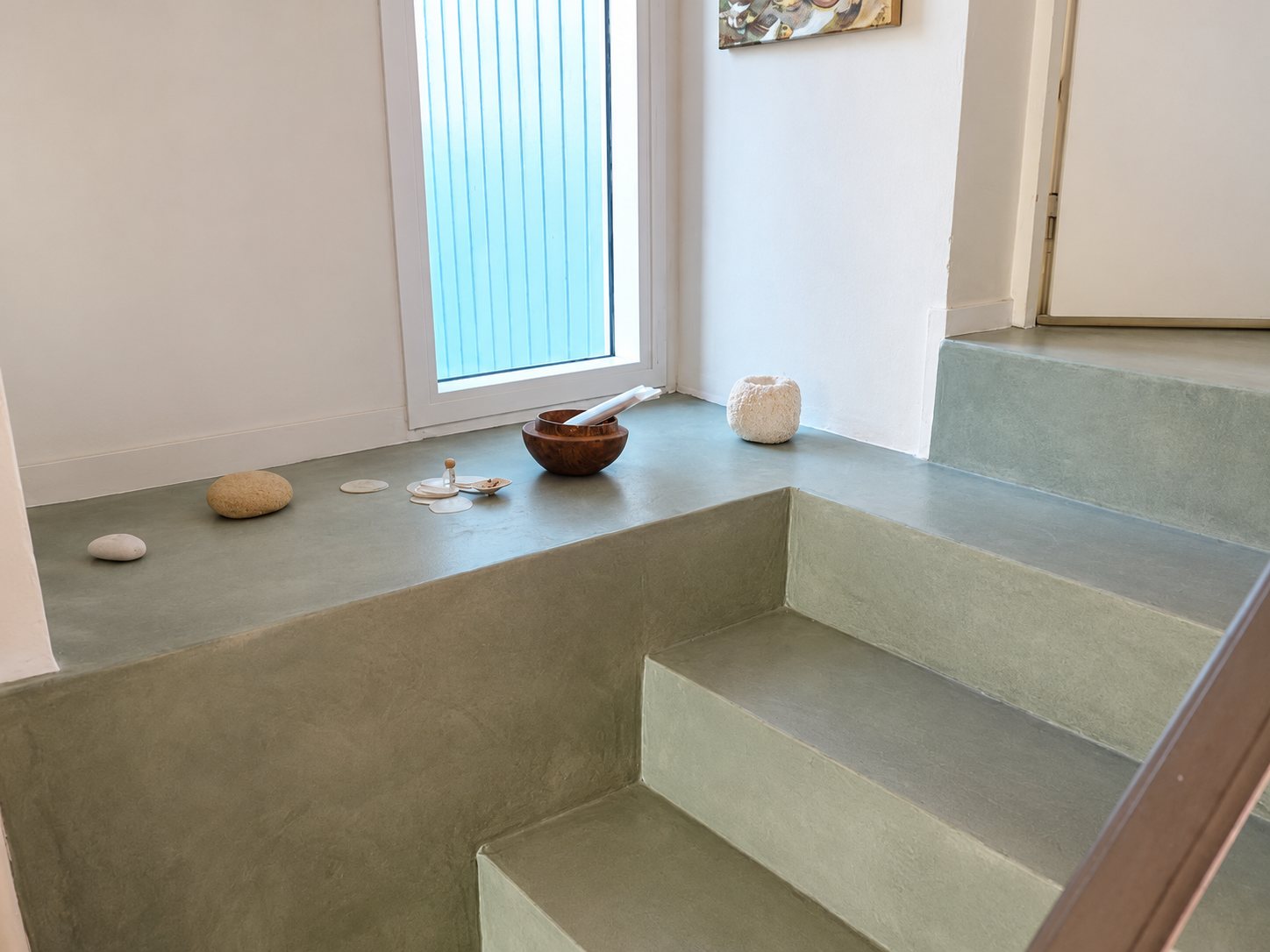 Un escalier en béton ciré vert-gris mène à une plateforme près d’une fenêtre, où sont posés des galets, un bol en bois et une bougie blanche.