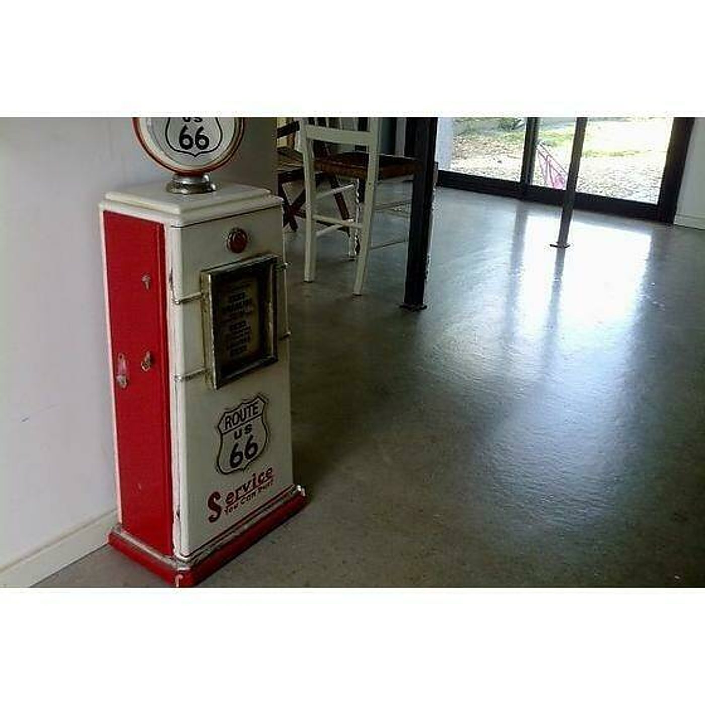 Un meuble décoratif rouge et blanc imitant une ancienne pompe à essence Route 66, posé dans une pièce lumineuse au sol gris.