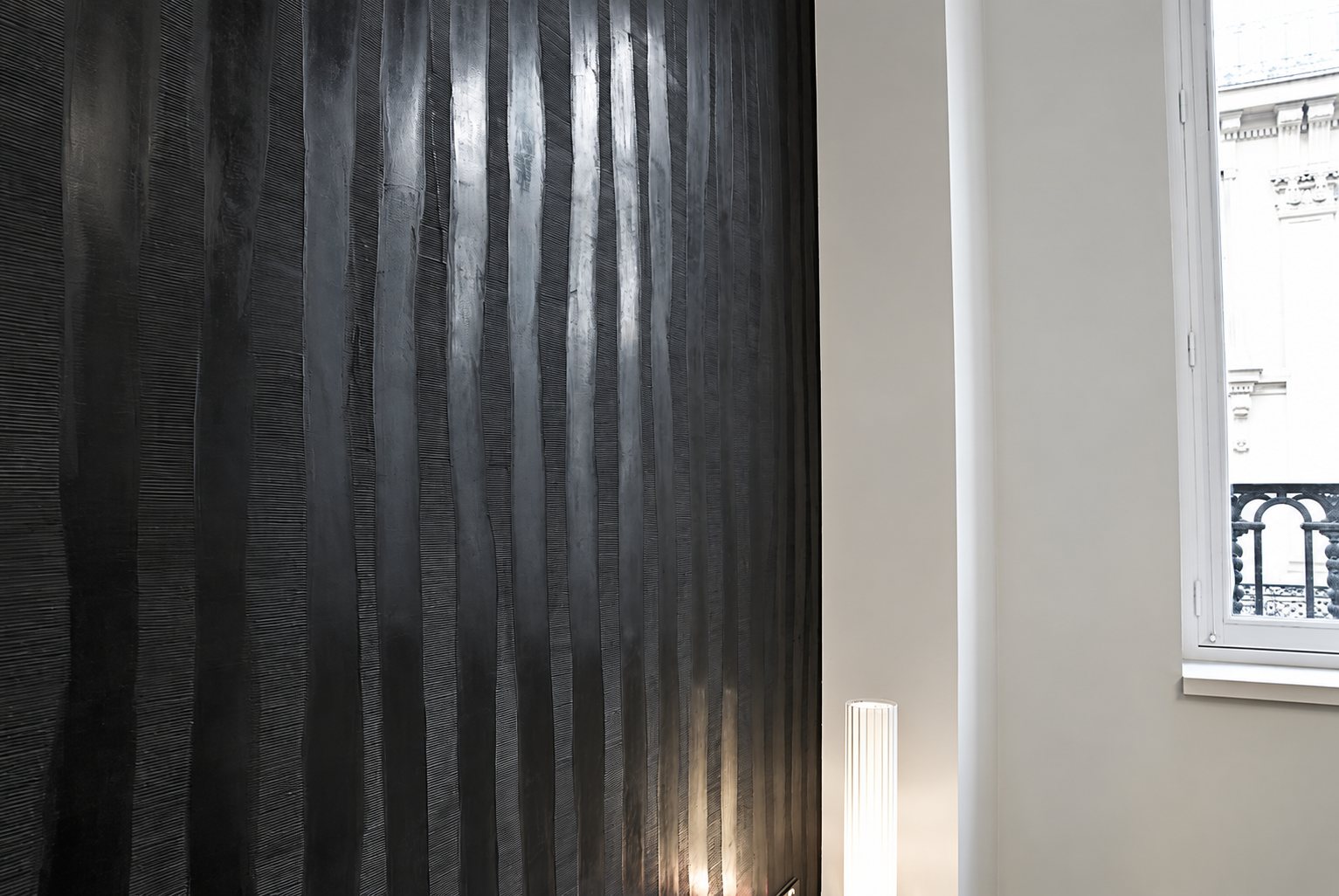 Un grand mur noir aux fines rayures verticales en relief, à côté d’un mur clair et d’une fenêtre donnant sur un immeuble haussmannien.