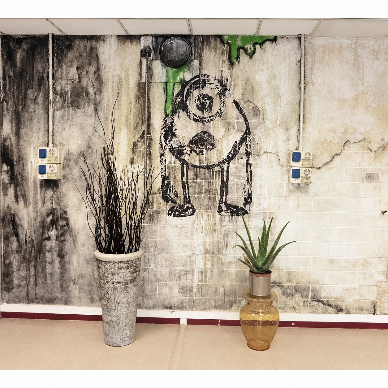 Un mur en briques vieilli orné d’un grand dessin noir d’un personnage à un œil, devant lequel sont posés un haut vase gris avec des branches sèches et un vase en verre ambré avec une plante d’aloès.