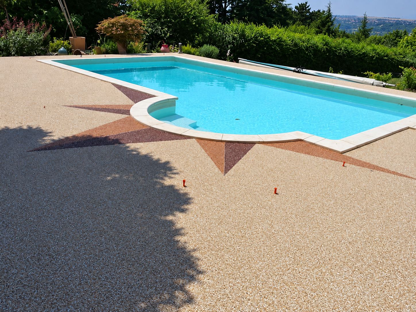 Piscine rectangulaire aux bords arrondis entourée d’une plage en revêtement de graviers beige, ornée d’une rose des vents rouge brun, dans un jardin avec vue dégagée.