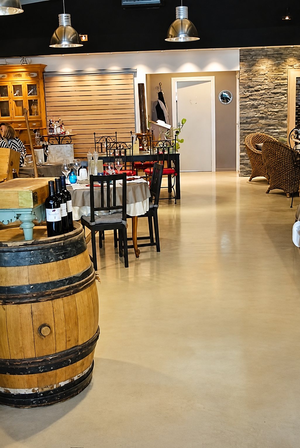 Intérieur lumineux d’un restaurant avec un tonneau en chêne portant des bouteilles de vin au premier plan, des tables dressées et des fauteuils en rotin au fond.