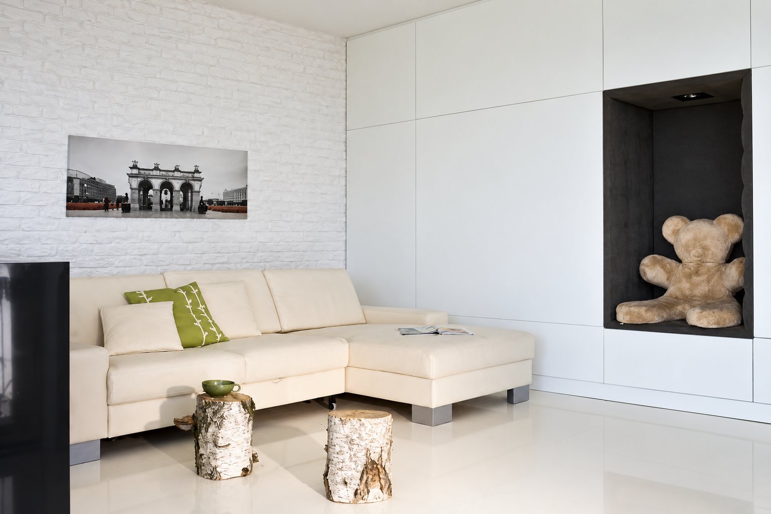 Salon lumineux tout en blanc avec un canapé d’angle crème, deux tabourets en bouleau, un panoramique en noir et blanc au mur et un gros ours en peluche dans une niche.
