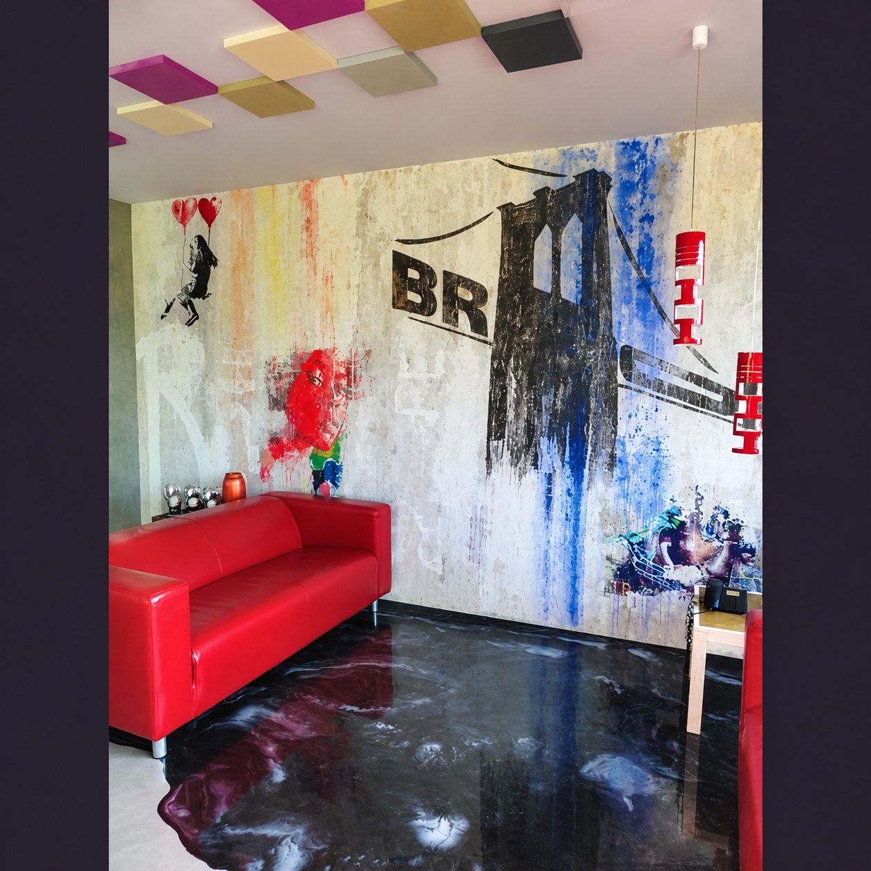 Un salon au mur orné d’une fresque de pont urbain en street art, avec un canapé en cuir rouge et un sol résiné sombre brillant.