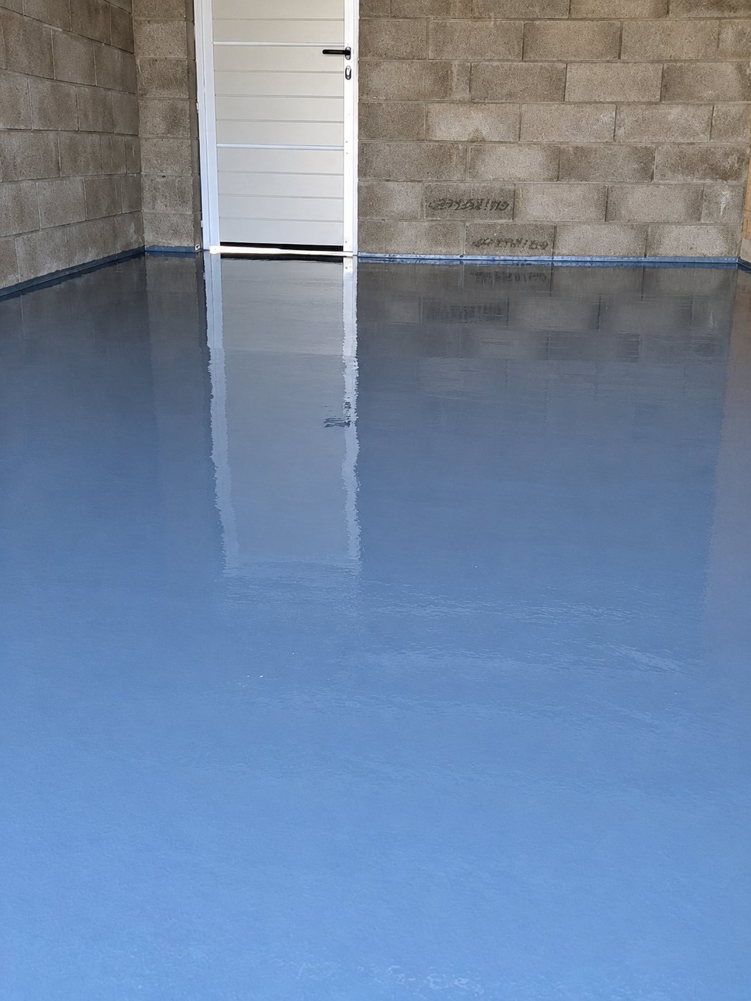 Un garage aux murs en parpaings dont le sol fraîchement recouvert de résine bleu-gris brillante reflète la porte blanche au fond.
