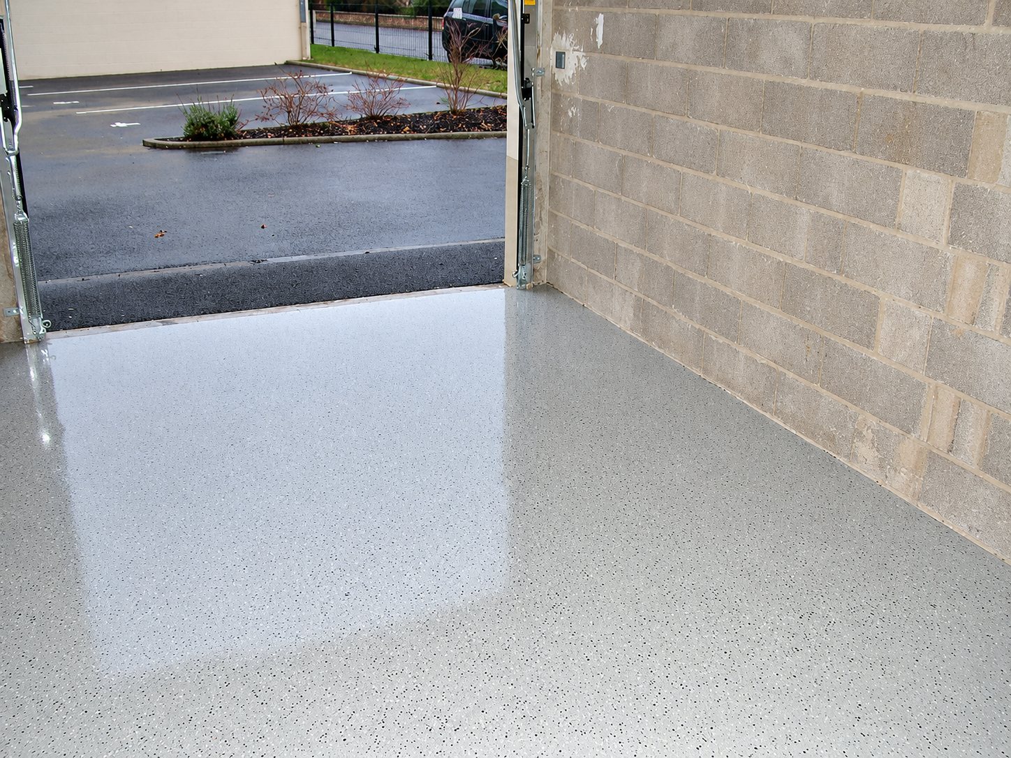 Intérieur d’un garage dont le sol est recouvert d’une résine grise brillante à paillettes, avec un mur en parpaings et un parking visible à travers la porte ouverte.