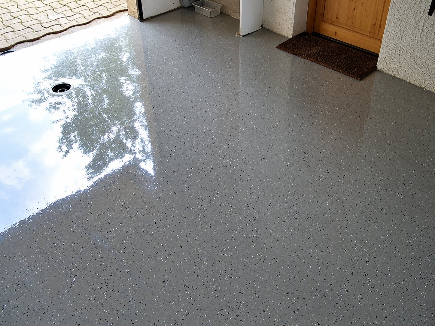Sol de garage revêtu d’une résine grise brillante parsemée de paillettes, avec un reflet de lumière, une évacuation et une porte en bois au fond.