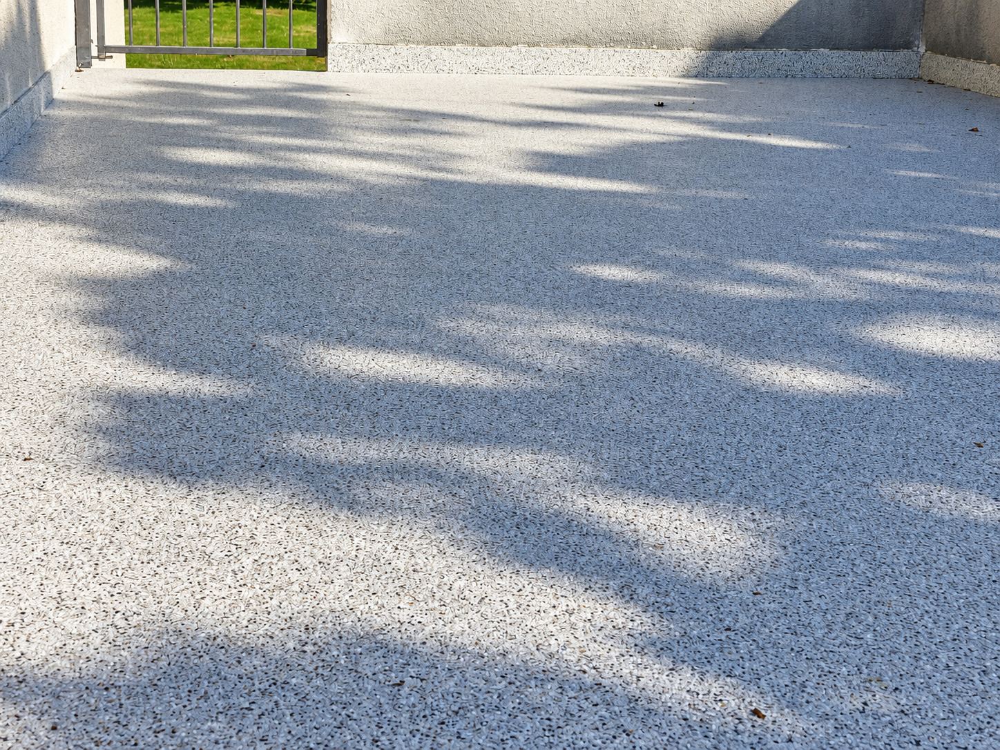 Une terrasse extérieure au revêtement de résine granulée gris clair, parcourue par les ombres d’un feuillage, avec un portillon métallique et de l’herbe au fond.