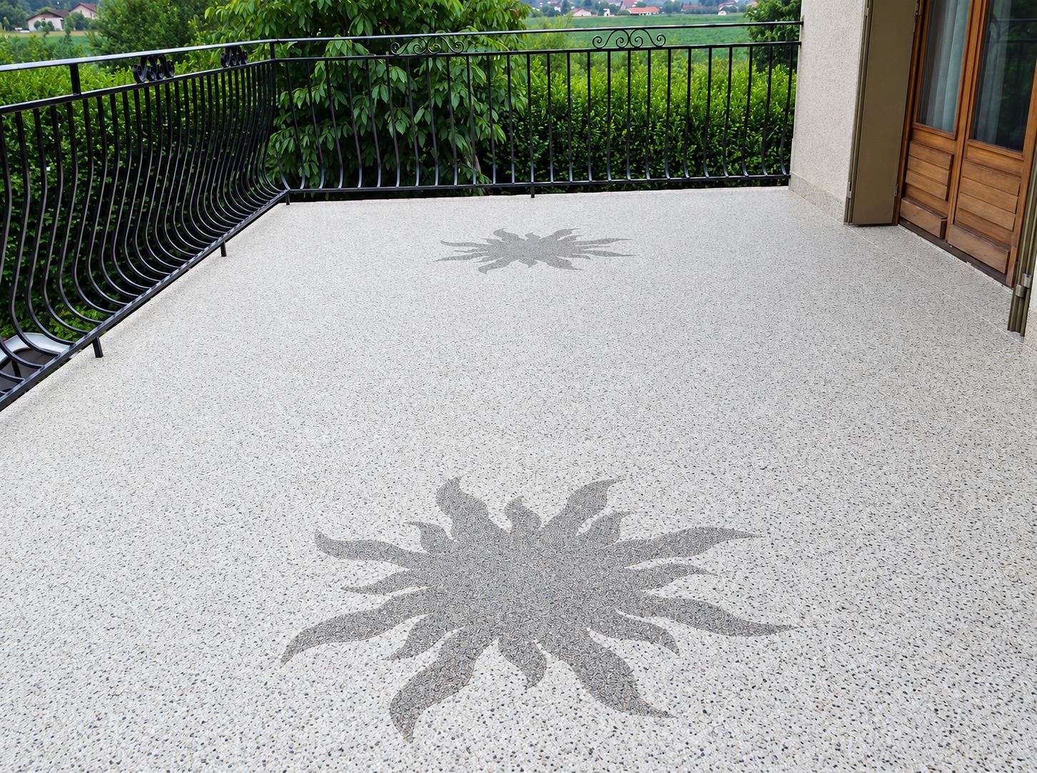 Une terrasse extérieure au sol en revêtement de résine beige moucheté, orné de deux motifs de soleil gris, bordée d’une balustrade en fer forgé noir.