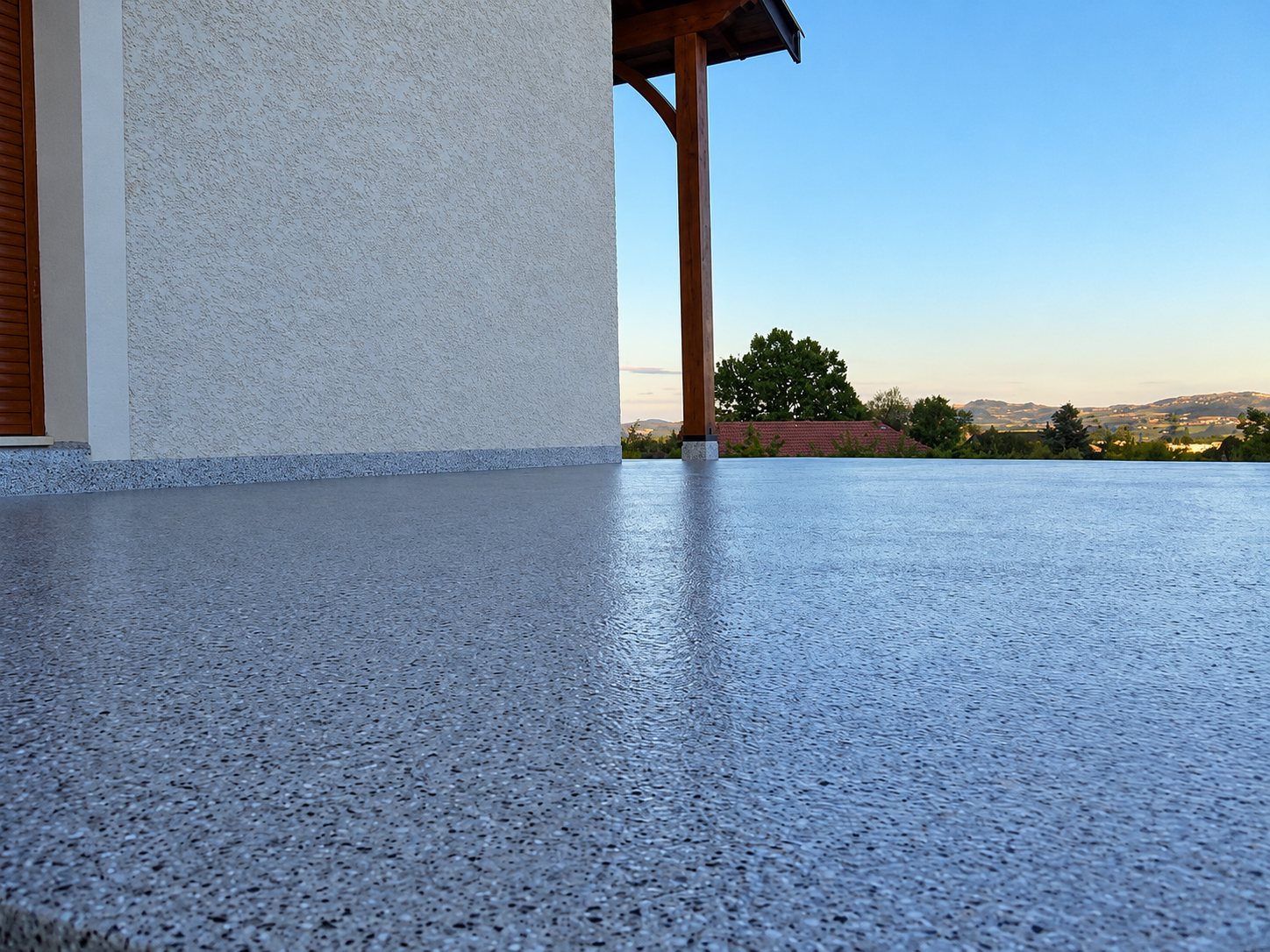 Vue au ras du sol d’une terrasse recouverte d’un revêtement gris granuleux et brillant, le long d’un mur crépi, avec un poteau en bois et un paysage de collines au loin.